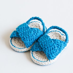 May include: A pair of crocheted baby sandals in blue and white. The sandals have a simple design with a strap across the top.