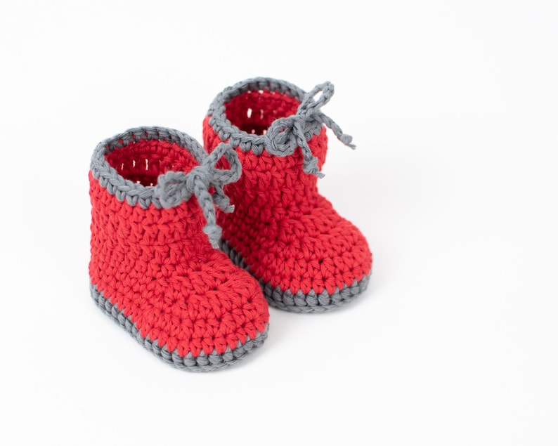 Op de afbeelding: Een paar rode gehaakte babyschoentjes met grijze afwerking en veters.