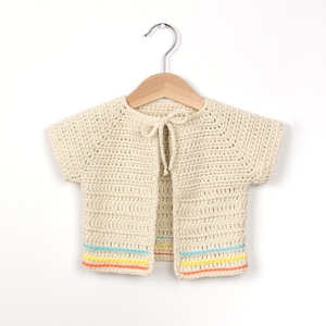 May include: A cream-coloured crocheted cardigan with short sleeves and a tie closure. The cardigan has a colourful trim with blue, yellow, and orange stripes.