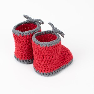 Op de afbeelding: Een paar rode gehaakte babyschoentjes met grijze bies. De schoentjes hebben een simpel ontwerp en zijn perfect voor een pasgeboren baby.
