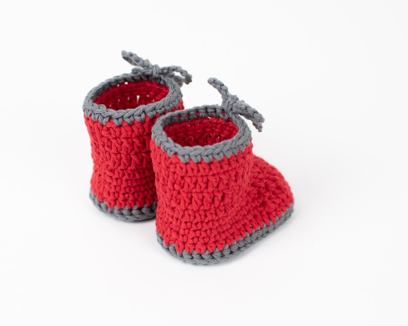 Op de afbeelding: Een paar rode gehaakte babyschoentjes met grijze bies. De schoentjes hebben een simpel ontwerp en zijn perfect voor een pasgeboren baby.