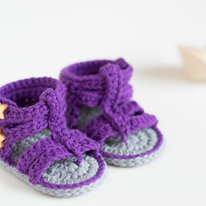 May include: A pair of purple crocheted baby sandals with gray soles and wooden star buttons.