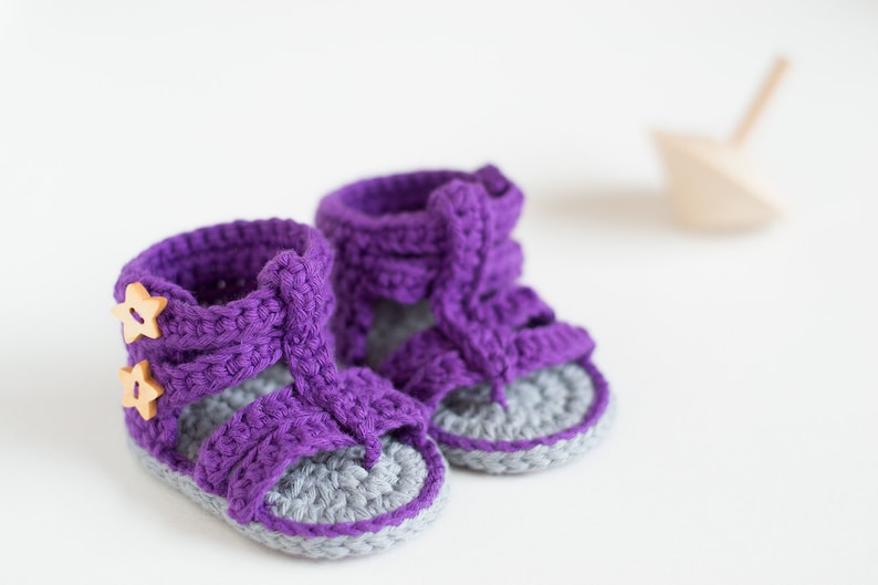 May include: A pair of purple crocheted baby sandals with gray soles and wooden star buttons.