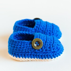 May include: A pair of blue crocheted baby shoes with a white trim and a brown button on each shoe.