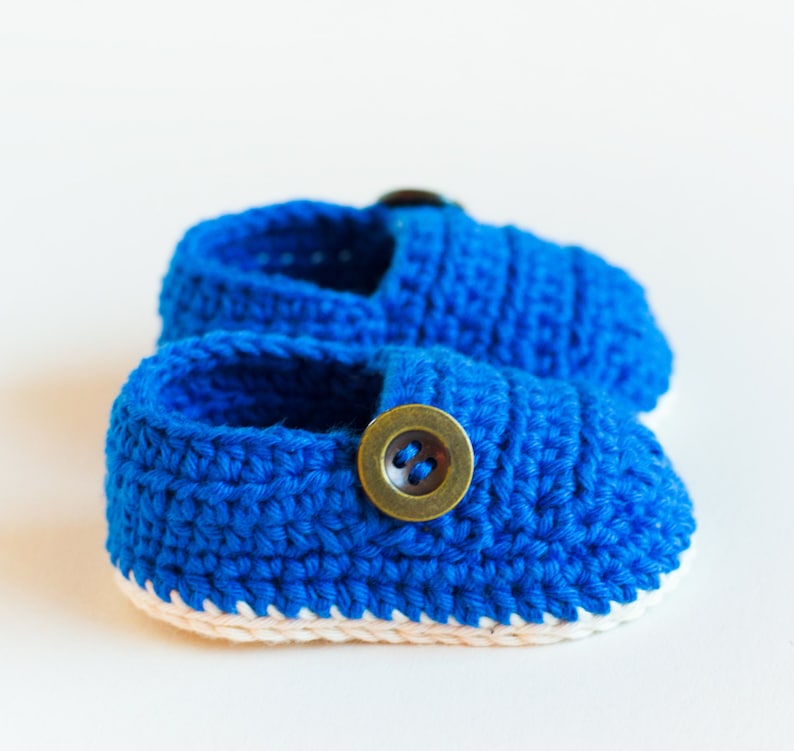May include: A pair of blue crocheted baby shoes with a white trim and a brown button on each shoe.