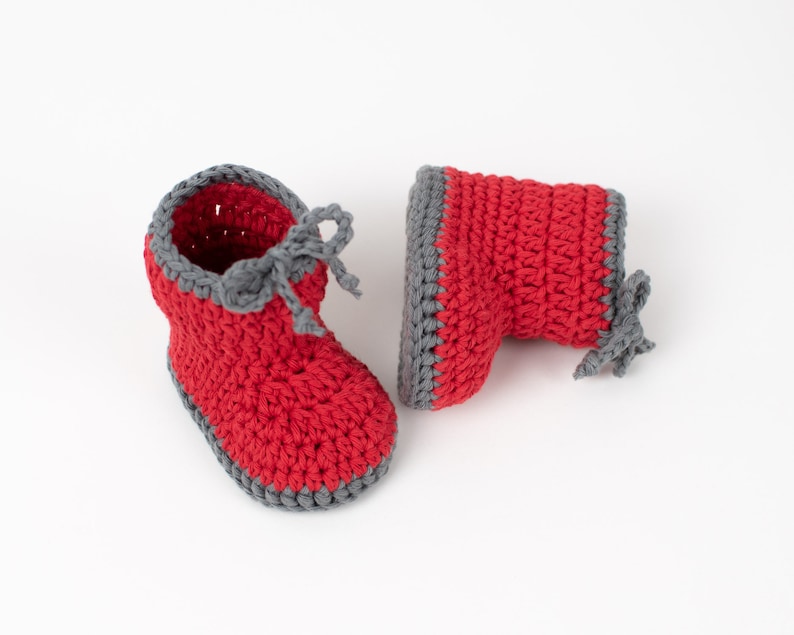 Op de afbeelding: Een paar rode gehaakte babyschoentjes met grijze afwerking. De schoentjes hebben een simpel ontwerp en zijn perfect voor een pasgeborene.