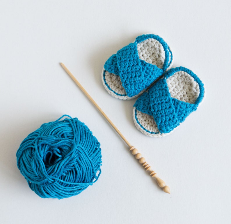 May include: Two crocheted baby sandals in blue and white with a ball of blue yarn and a wooden knitting needle.