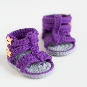 May include: A pair of purple crocheted baby sandals with gray soles and wooden star buttons.