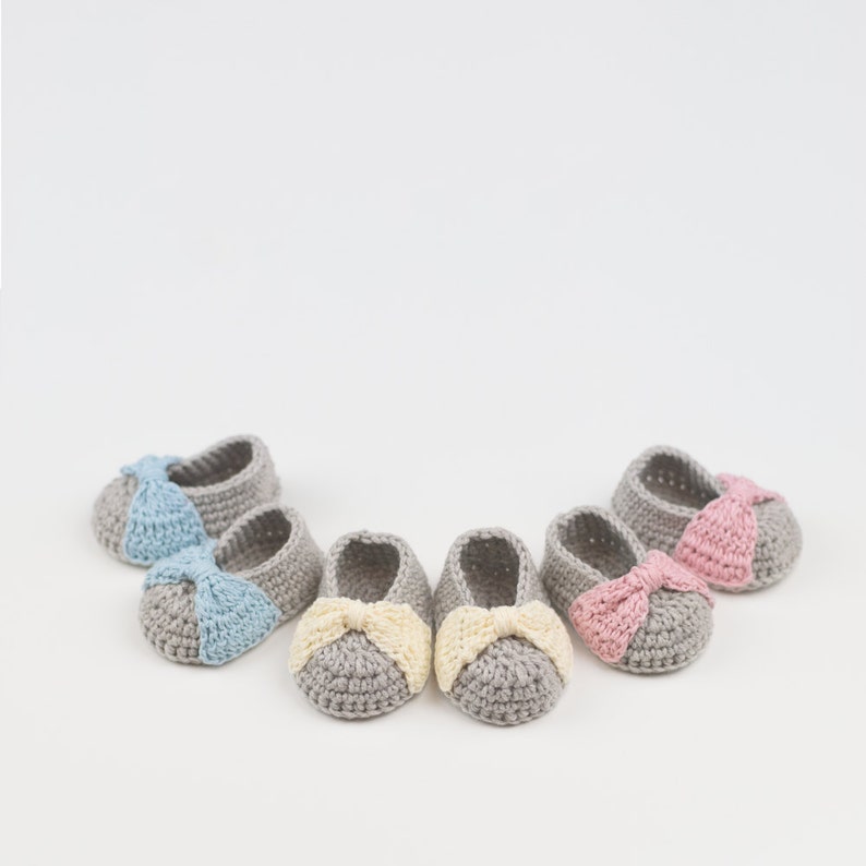 Puede incluir: Cinco pares de botines de beb&eacute; tejidos a crochet con lazos de diferentes colores. Los botines son grises con un lazo azul claro, rosa o amarillo.