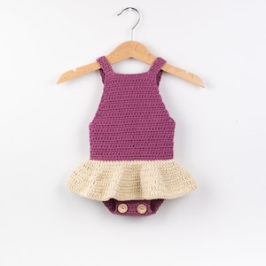 May include: A crocheted baby romper with a purple bodice and a cream-coloured ruffle skirt. The romper has two wooden buttons at the crotch.