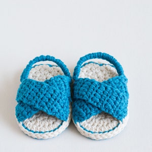 May include: A pair of crocheted baby sandals in blue and white. The sandals have a criss-cross strap design and a white sole.