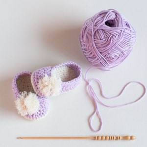 May include: Two lavender crocheted baby booties with white pom-poms, a ball of lavender yarn, and a wooden crochet hook.
