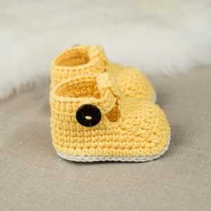 May include: A pair of yellow crocheted baby shoes with a white trim and a brown button on the strap.