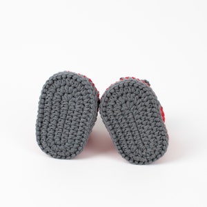 Op de afbeelding: Een paar grijze gehaakte babyschoentjes met rode bies. De schoentjes worden van onderaf getoond, met de gehaakte zolen zichtbaar.