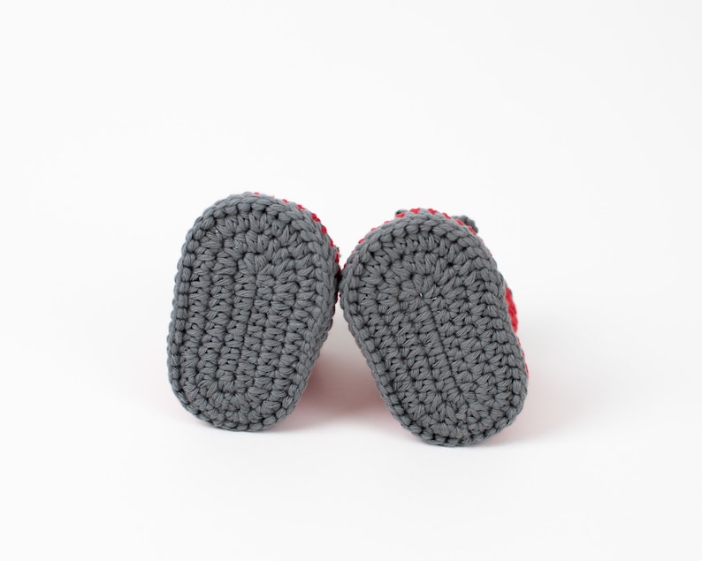 Op de afbeelding: Een paar grijze gehaakte babyschoentjes met rode bies. De schoentjes worden van onderaf getoond, met de gehaakte zolen zichtbaar.