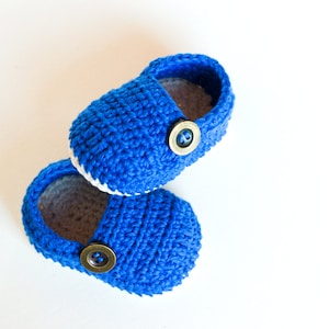 Può includere: Un paio di scarpine per neonati in crochet blu con bordi bianchi e bottoni marroni.