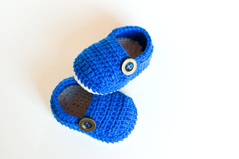 May include: A pair of blue crocheted baby shoes with white trim and brown buttons.