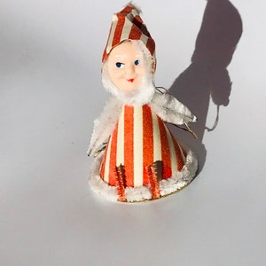 May include: A vintage Christmas ornament shaped like a person wearing a red and white striped hat and dress. The ornament has a white fuzzy trim and a painted face with blue eyes.