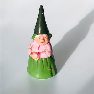 May include: A green and pink ceramic gnome figurine with a green pointed hat. The gnome is smiling and has a rosy complexion.