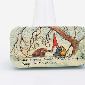 Op de afbeelding: Een vintage illustratie van een kabouter die kleine knaagdieren voert tijdens de lange, strenge winters. De afbeelding is gedrukt op een rechthoekige, blauw-witte blik.