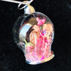 Könnte beinhalten: Eine silberne Halskette mit einem Glasanhänger, der getrocknete Blumen und einen rosa Kristall enthält. Der Anhänger ist rund und hat einen silbernen Rand.