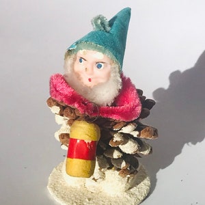 May include: A vintage Christmas bauble made from a pine cone, featuring a felt elf with a blue hat and pink collar, holding a small red and yellow lantern.