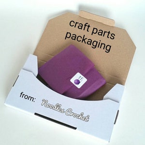 May include: A white box with the text "from: Noodles Crochet" on the bottom. The box is open and contains a purple tissue paper package with a small white sticker that says "Noodles Crochet". The box is labeled "craft parts packaging" on the top.