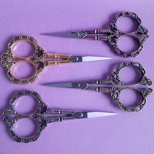 May include: Three pairs of decorative scissors with ornate handles. The scissors are gold, silver, and bronze in colour. The handles are decorated with floral designs.