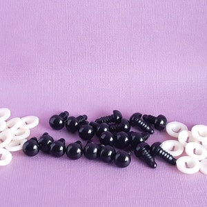 May include: A collection of black plastic safety eyes with screw backs and white plastic washers. The eyes are arranged in a pile on a purple surface.