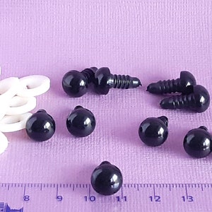 May include: A close-up of black plastic safety eyes with a screw back. The eyes are arranged on a purple surface with a ruler in the background. The ruler is marked in inches.