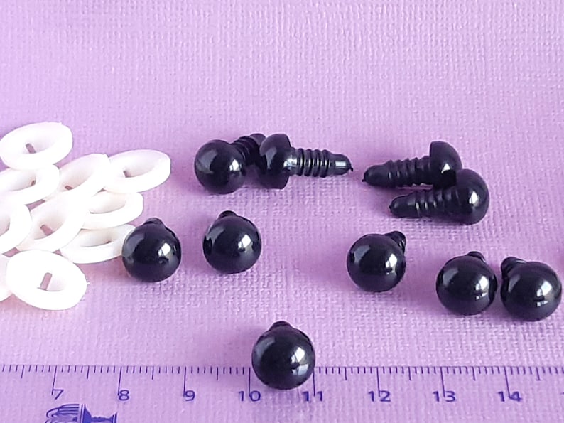 May include: A close-up of black plastic safety eyes with a screw back. The eyes are arranged on a purple surface with a ruler in the background. The ruler is marked in inches.