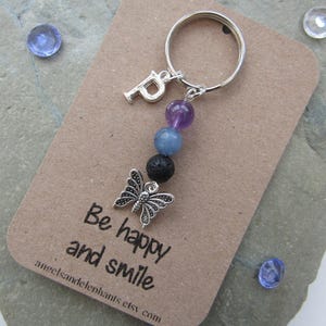 May include: A silver charm keyring with a butterfly charm, a letter "P" charm, and purple, blue, and black beads. The keyring is attached to a card that says "Be happy and smile".