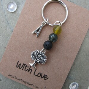 May include: A silver charm keyring with a tree charm, a letter A charm, and three beads: a yellow bead, a green bead, and a black bead. The keyring is on a brown card with the text "With Love" and a website address.