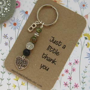 May include: A brown card with the text "Just a little thank you" and a silver charm keyring with a heart, tree, and letter B. The keyring is made of green and brown beads.