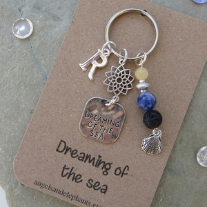 May include: A silver keyring with a charm that says "Dreaming of the Sea". The keyring also has a silver letter "R", a silver mandala charm, a yellow bead, a blue bead, a black lava bead, and a silver seashell charm.