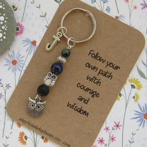 May include: A silver charm keychain with a silver owl charm, blue and green beads, and a silver letter J charm. The keychain is attached to a card with the text "Follow your own path with courage and wisdom".