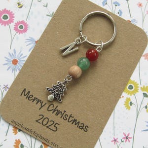 May include: A silver keyring with a decorative Christmas tree charm, letter "N" charm, and beads in red, green, and wood tones. The keyring is on a brown card with the text "Merry Christmas 2025".