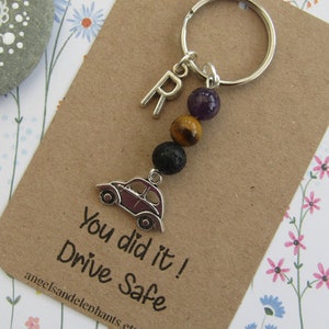 May include: A brown card with the text "You did it! Drive Safe" and a silver charm of a car attached to a silver key ring with a purple amethyst bead, a tiger's eye bead, and a black lava bead. The charm is attached to a silver letter "R".