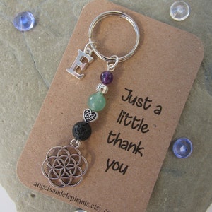 May include: A silver keyring with a charm featuring a silver letter E, a purple amethyst bead, green aventurine beads, a silver heart charm, a black lava bead, and a silver flower of life charm. The keyring is attached to a brown card with the text "Just a little thank you".