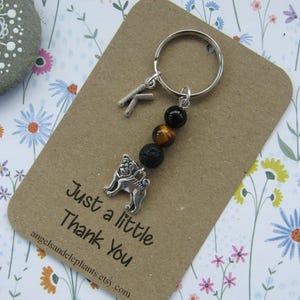 May include: A silver pug charm keyring with black lava rock and tiger's eye beads. The keyring is attached to a silver key ring. The keyring is on a brown card with the text "Just a little Thank You" and the website address "angelsandelephants.etsy.com".
