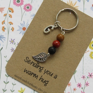 May include: A keyring featuring a silver letter 'G' charm, wooden beads in brown, red, and black, and a leaf charm. The keyring is attached to a brown card with the text 'Sending you a warm hug'. The background has a floral pattern.