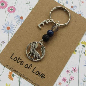 May include: A silver-coloured keyring featuring a wolf howling at the moon pendant. The keyring includes a letter "E" charm, and beads in shades of grey, blue, and black. The keyring is displayed on a brown card with the text "Lots of Love".