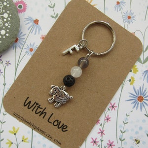 May include: A silver charm keychain with a small silver letter F, three beads, and a silver charm of a sheep. The beads are black, white, and grey. The keychain is on a brown card with the text "With Love" and a website address.