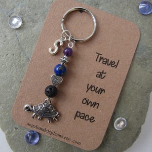 May include: A silver keyring with a turtle charm, letter "S" charm, and beads in purple, blue, and black. The keyring is on a brown card with the text "Travel at your own pace."