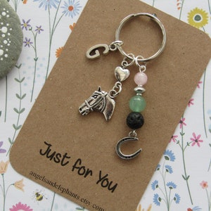 May include: A silver horse head charm, a horseshoe charm, a silver heart charm, a pink heart charm, a pink gemstone bead, a green gemstone bead, a black lava bead, and a silver letter G charm are attached to a silver key ring. The charms are hanging from a brown card with the text "Just for You" and the website address "angelsandelephants.etsy.com".