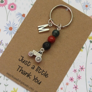 May include: A silver tractor charm keyring with a letter "M" charm and three beads in red, green, and black. The keyring is attached to a brown card with the text "Just a little Thank You". The background has a floral pattern.
