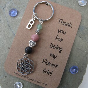 May include: A silver charm keychain with a flower of life charm, lava rock beads, a pink heart charm, a pink stone bead, a green stone bead, and a silver letter "B" charm. The keychain is attached to a brown card with the text "Thank you for being my Flower Girl".