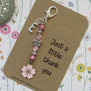 May include: Pink and silver charm with a butterfly, flower, and the letter E. The charm is attached to a silver keyring. The charm is on a brown card with the text "Just a little thank you".