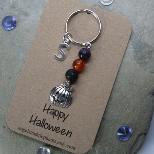 May include: A silver charm keychain with a pumpkin charm, black lava beads, orange and red beads, and a silver letter S charm. The keychain is on a brown card with the text "Happy Halloween" and a website address.