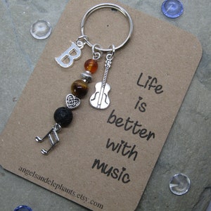 May include: A silver keyring with a charm that says "Life is better with music". The charm features a silver guitar, a silver music note, a lava rock bead, a tiger's eye bead, a heart charm, and a silver letter "B".